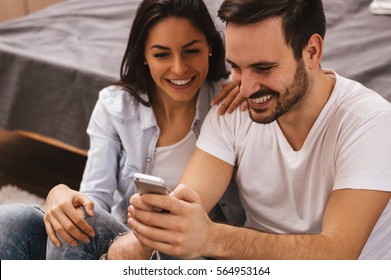 Cheerful Couple Laughing Watching Media In Smart Phone Sitting On Floor.