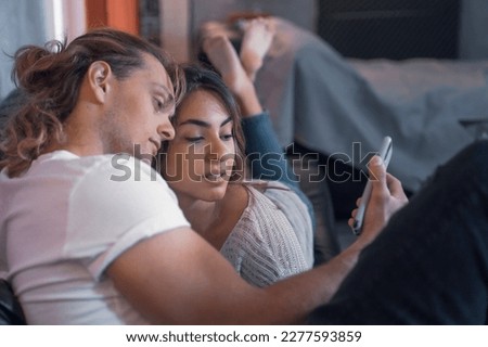 Cheerful couple of romantic serious caucasian guys sharing a night message in living room searching and messaging watching their cellphone device. Male fun guy enjoyng his life looking a smart girl