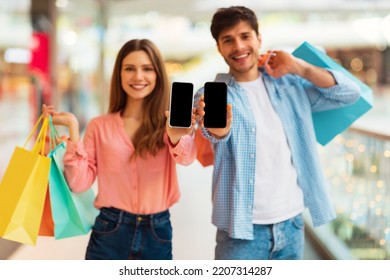 Cheerful Couple Showing Smartphone Screen Recommending Mobile Shopping Application Posing With Colorful Shopper Bags In Modern Hypermarket. Selective Focus On Phones. Mockup