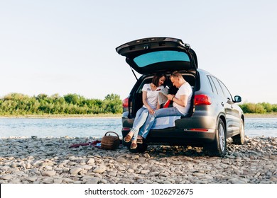 Cheerful couple sitting in car open trunk and watching map, roadtrip concept