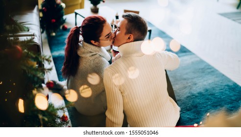 Cheerful couple sitting on floor kissing with tenderness with glasses of wine in hands in decorated living room celebrating Christmas