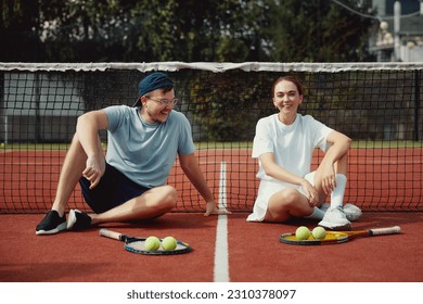 Cheerful couple sitting on the tennis court floor with rackets and ball. Friends are Smiling and have fun. Sports active game with friends. Tennis players after match - Powered by Shutterstock