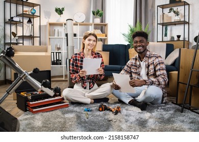 Cheerful Couple Sitting Together At Living Room And Reading Furniture Assembly Instructions At New Home. Multi Ethnic Family Looking Forward To New Stage In Their Common Life.