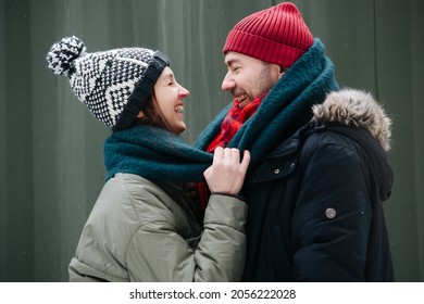 Cheerful couple standing in front of each other outdoors at winter. Both wearing hats, jackets and scarfs. In front of corrugated sheet house wall.