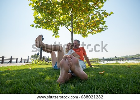 Cheerful couple taking selfie and having fun while sitting on a green meadow barefoot, under tree. Sunny morning in the park. Wide angle, back light.