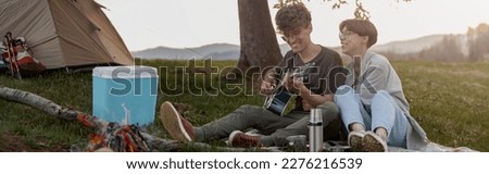 Cheerful couple of travelers sitting at fire in camp and man playing on guitar. Travel life