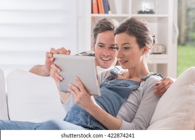 Cheerful Couple Using A Tablet At Home, The Young Woman Is Pregnant, She Si Lying On A White Couch And His Husband Look The Tablet Over Her Shoulder