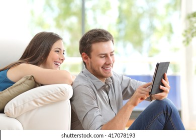 Cheerful couple using a tablet on line sitting in the living room at home with a window in the background - Powered by Shutterstock