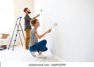 Cheerful couple young  man and woman smiling and  painting white wall with roller during renovation in new apartment