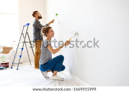 Cheerful couple young  man and woman smiling and  painting white wall with roller during renovation in new apartment
