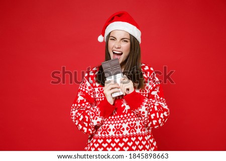 Cheerful crazy young Santa woman 20s wearing sweater Christmas hat biting eating chocolate bar isolated on bright red wall background studio portrait. Happy New Year celebration merry holiday concept