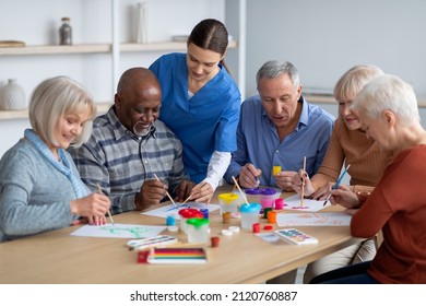 Cheerful Creative Multiracial Senior Men And Women Sitting Around Table And Drawing At Nursing Home, Doing Arts And Crafts Together, Young Lady Nurse Assisting Group Of Elderly People, Copy Space