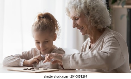 Cheerful curious adorable small kid girl learning playing checkers with caring smiling old senior grandmother, involved in interesting wooden boardgame, improving strategic skills together at home.