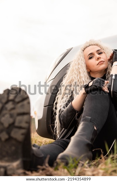 Cheerful curly blonde long hair rock woman\
laugh sitting on dry grass near automobile wheel relaxing enjoy\
travel journey. Happy fashion female resting on highway countryside\
scenery autumn\
landscape