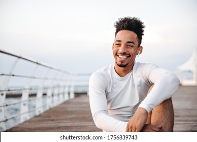 Cheerful curly dark-skinned man in white long-sleeved t-shirt smiles sincerely and rests near sea. Sportsman listens to music in wireless headphones.