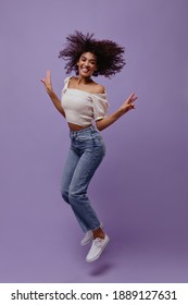 Cheerful curly girl jumps on purple background. Attractive dark-skinned woman in jeans and white top shows peace signs on isolated.