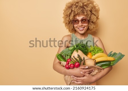 Cheerful curly haired woman carries paper bag full of fresh vegetables and fruits picked up from home garden wears sunglasses and t shirt isolated over beige background blank space for advertisement