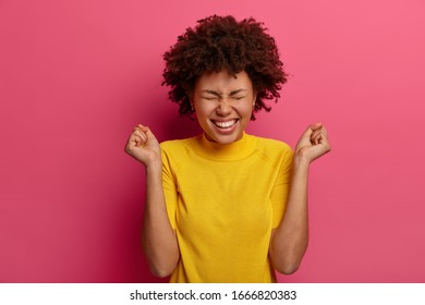 Cheerful curly haired woman clenches fists with positive expression, rejoices wonderful news, smiles happily, squints face with pleasure, wears yellow clothes, stands against pink vibrant background