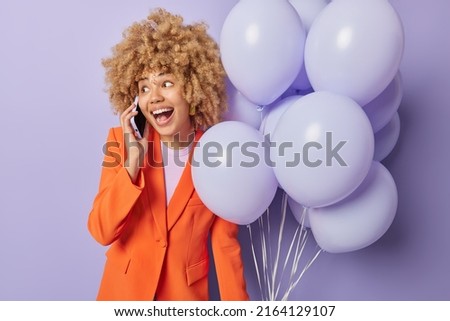 Cheerful curly haired woman dressed in fashionable clothes makes phone call holds bunch of inflated ballons invites friends on party isolated over purple background. Festive occasion concept