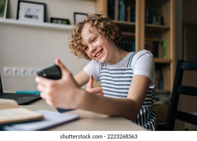 Cheerful Curly Little Girl Holding And Using Mobile Phone Showing Thumb Up, Browsing Social Network Apps, Surfing Internet And Scrolling News Feed, Reading Electronic E-book At Table With Laptop.