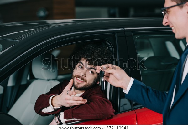 cheerful curly man sitting in red car and looking\
at key in hand of car dealer\
