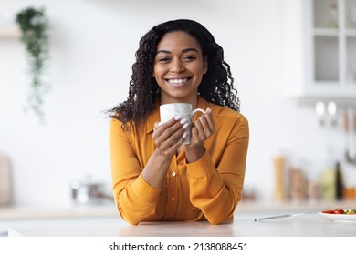 Cheerful curly millennial african american lady drinking tea at home, sitting at table at kicthen, holding coffee mug and smiling at camera, having coffee break while working from home, copy space - Powered by Shutterstock