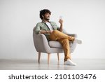 Cheerful curly young indian man chatting with girlfriend while relaxing in armchair at home, using modern mobile phone, checking social media, using mobile app, panorama with copy space