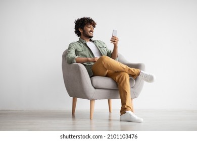 Cheerful curly young indian man chatting with girlfriend while relaxing in armchair at home, using modern mobile phone, checking social media, using mobile app, panorama with copy space