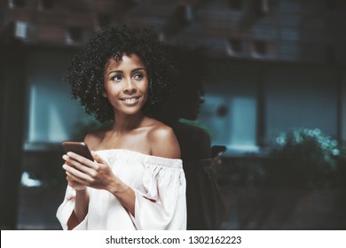 A cheerful curly-haired young Brazilian woman is leaning against a mirror wall and looking aside while using the smartphone to talk with her colleague, with copy space zone on the right for your text - Powered by Shutterstock