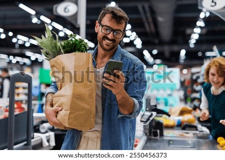 Cheerful customer is using a smartphone while holding a brown paper bag full of fresh groceries at the checkout counter of a modern supermarket, with a friendly cashier in the background