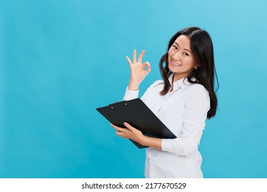 Cheerful Cute Asian Businesswoman In Classic Office Dress Code Holds Folder Tablet Show Okay Gesture Posing Isolated On Over Blue Studio Background. Cool Business Offer. Job Interview Concept