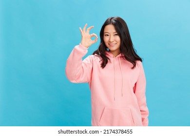 Cheerful cute Asian student young lady in pink hoodie sweatshirt show Okay sign gesture posing isolated on over blue studio background. The best offer for ad. People Emotions for Everyday concept - Powered by Shutterstock