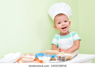 Cheerful cute baby boy in a chef costume laughs standing at the table where doing cookies.	
