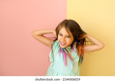 Cheerful Cute Funny Girl With Bright Multi Colored Strands Of Hair Laughs Merrily And Straightening Hair With Hands. Child With Artificial Locks Of Hair On A Yellow-pink Studio Background. 