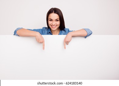 Cheerful cute girl is standing behind the white blank banner and pointing down at a copyspace - Shutterstock ID 624183140