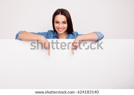 Cheerful cute girl is standing behind the white blank banner and pointing down at a copyspace