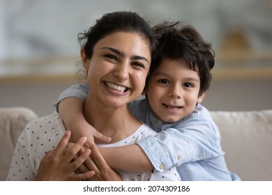 Cheerful Cute Indian Preschooler Kid Embracing Happy Mother On Couch, Looking At Camera, Smiling. Young Mom And Kid Hugging With Love, Affection, Tenderness. Head Shot Home Portrait