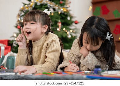 A cheerful and cute young Asian girl is enjoying playing and drawing a Christmas card with her sister while lying on the living room's floor together. Merry Christmas - Powered by Shutterstock