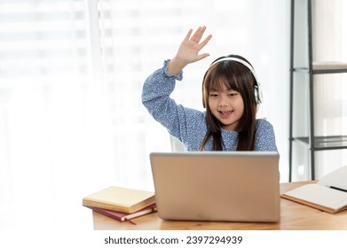 A cheerful and cute young Asian girl is raising her hand to answer a question while studying online with her private tutor indoors. Kid and online learning concepts - Powered by Shutterstock