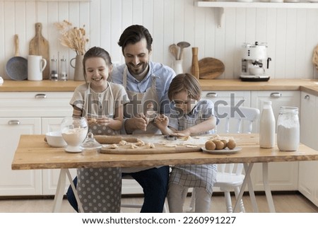 Cheerful dad teaching positive little children to bake, shaping dumplings over floury table, with dough, eggs, milk, smiling, laughing, enjoying culinary hobby, family leisure