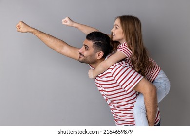 Cheerful daddy piggybacking daughter, making flying superhero hand, fist forward gesture, little girl riding fathers back, playing active game together. Indoor studio shot isolated on gray background. - Powered by Shutterstock