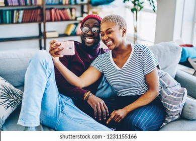 Cheerful dark skinned couple in love resting at home interior posing for selfie on smartphone camera,happy young hipsters making image on modern mobile phone laughing at living room together