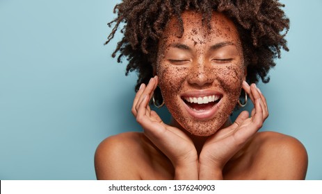 Cheerful dark skinned lady applies coffee scrub on face, pampers skin, closes eyes from pleasure, smiles positively has bare shoulders, stands against blue background, free space aside. Beauty concept