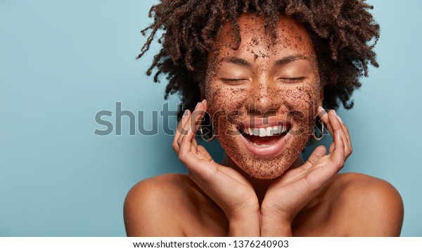 Cheerful dark skinned lady applies coffee scrub\
on face, pampers skin, closes eyes from pleasure, smiles positively\
has bare shoulders, stands against blue background, free space\
aside. Beauty concept