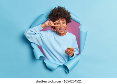 Cheerful dark skinned woman with Afro hair shows peace gesture over eye holds mobile phone dressed in casual clothing poses through torn paper hole of blue background. Body language concept. - Powered by Shutterstock