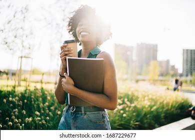 Cheerful dark skinned woman with curly hair laughing feeling carefree during free time outdoors, smiling african american hipster girl enjoying sunny day in summer rest with coffee to go in park