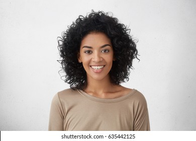 Cheerful dark-skinned girl smiling broadly, rejoicing at her victory in competition among young writers, standing isolated against grey wall background. People, success, youth and happiness concept