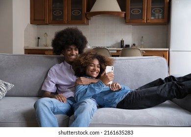 Cheerful Dating Teen Black Couple Using Online App On Smartphone At Home. Happy Young African Girlfriend And Boyfriend Relaxing On Couch, Laughing, Smiling, Shopping On Internet On Mobile Phone