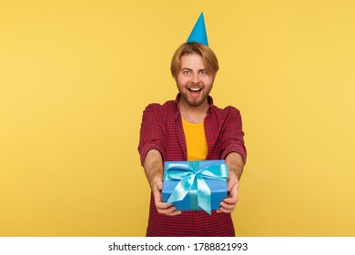 Cheerful delighted guy in funny party cone giving gift box, looking at camera with toothy smile, greeting happy birthday, congratulating on anniversary. studio shot isolated on yellow background