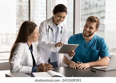 Cheerful diverse clinic medical colleagues in uniforms using tablet computer at workplace together, talking, laughing, discussing healthcare online service, enjoying modern Internet technology - Powered by Shutterstock